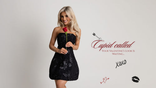 Woman standing against white backdrop in a studio wearing a strapless black mini dress holding a red rose for a valentine's photoshoot.