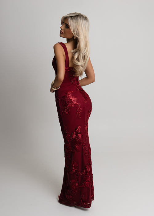 Woman standing against a light backdrop in a studio wearing a wine colour maxi dress with 3-D embroidery applique flowers, square neckline and thigh high slit, posed with hand on hip. 