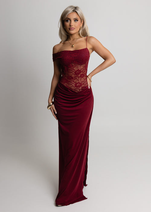 Woman standing against light backdrop in a studio, wearing a burgundy lace maxi dress with Lycra fitting maxi skirt, paired with gold jewellery and posed with one hand on hip.