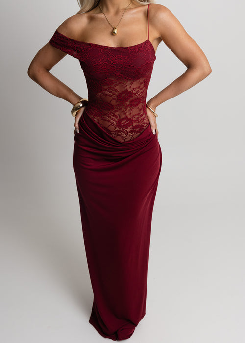 Close up of woman standing against light backdrop in a studio, wearing a burgundy lace maxi dress with Lycra fitting maxi skirt, paired with gold jewellery and posed with both hands on hip.