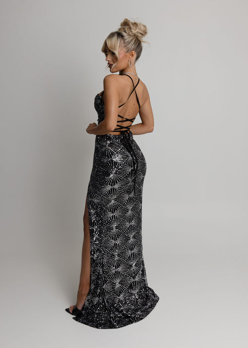 Women's black maxi dress embellished with silver glitter patterns all over fabric. Spaghetti straps and thigh high leg slit. Model is 5ft2' and wears a size 6.