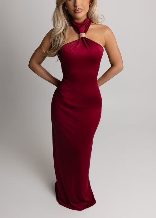 Woman standing in a studio against a light background, wearing a red jersey maxi dress with halter neck design, and gold jewellery, posing with one hand on her hip.