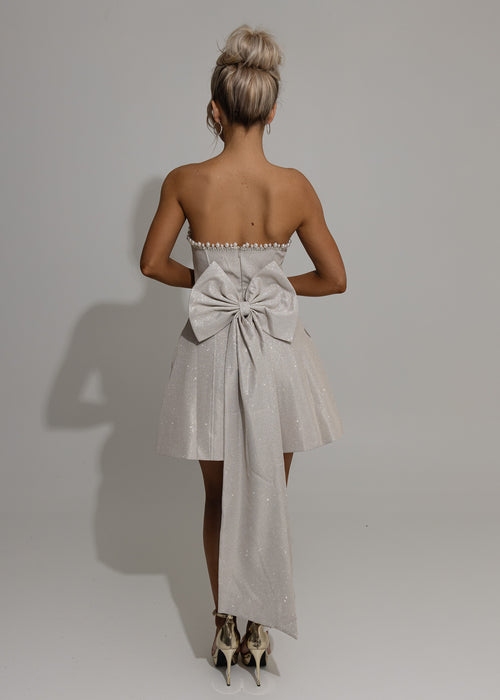 Women's champagne glitter mini dress with large detachable bow on the back of mini dress. Pearl and diamante trim embellishments with a strapless fit. Model is 5ft2' and wears a size XS 6.