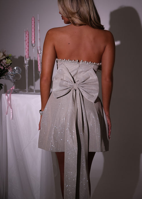 Women's champagne glitter mini dress with large detachable bow on the back of mini dress. Pearl and diamante trim embellishments with a strapless fit. Model is 5ft2' and wears a size XS 6.