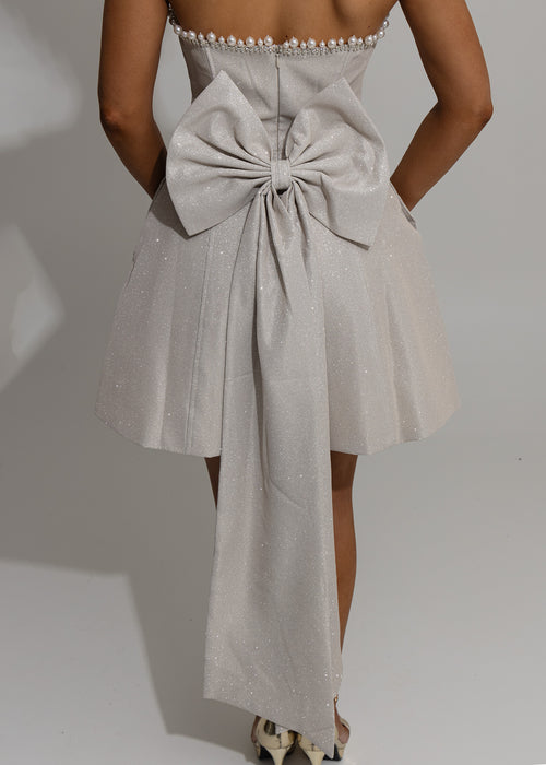 Women's champagne glitter mini dress with large detachable bow on the back of mini dress. Pearl and diamante trim embellishments with a strapless fit. Model is 5ft2' and wears a size XS 6.