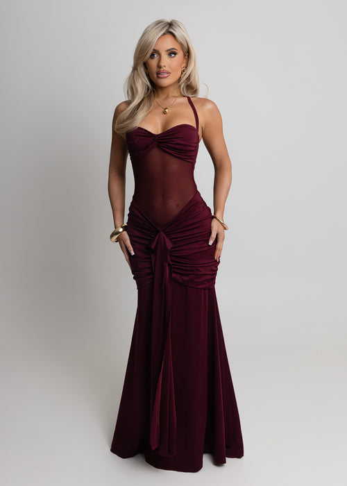 Woman standing against a light backdrop wearing a wine maxi dress with halter neckline, sheer mesh waist and ruched detailing from hips down to the floor, paired with gold jewellery and posing with hands on hips.