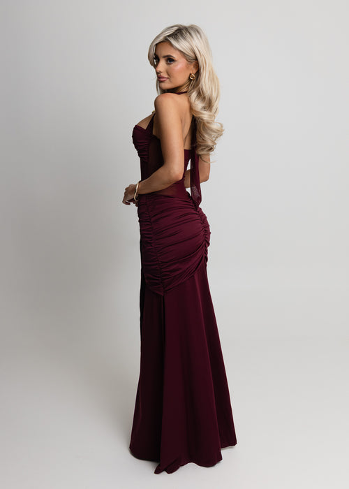 Woman standing against a light backdrop wearing a wine maxi dress with halter neckline, sheer mesh waist and ruched detailing from hips down to the floor, paired with gold jewellery and posing with hands on hips.