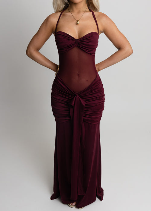 Woman standing against a light backdrop wearing a wine maxi dress with halter neckline, sheer mesh waist and ruched detailing from hips down to the floor, paired with gold jewellery and posing with hands on hips.