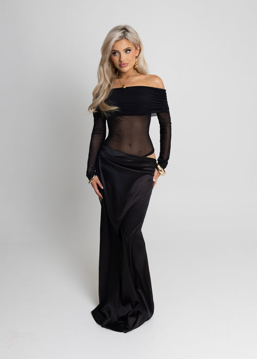 Woman standing against a light background wearing a black maxi dress with long mesh sleeves, cut out waist detail and Bardot style neckline, paired with gold jewellery and posed with hand on hip.
